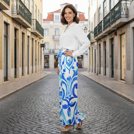 Full-body shot of a woman wearing Ondas Wide Leg Pants featuring a vibrant blue and white Mediterranean scroll print, paired with a white sleeveless top and straw purse, standing on a path at a tropical resort.