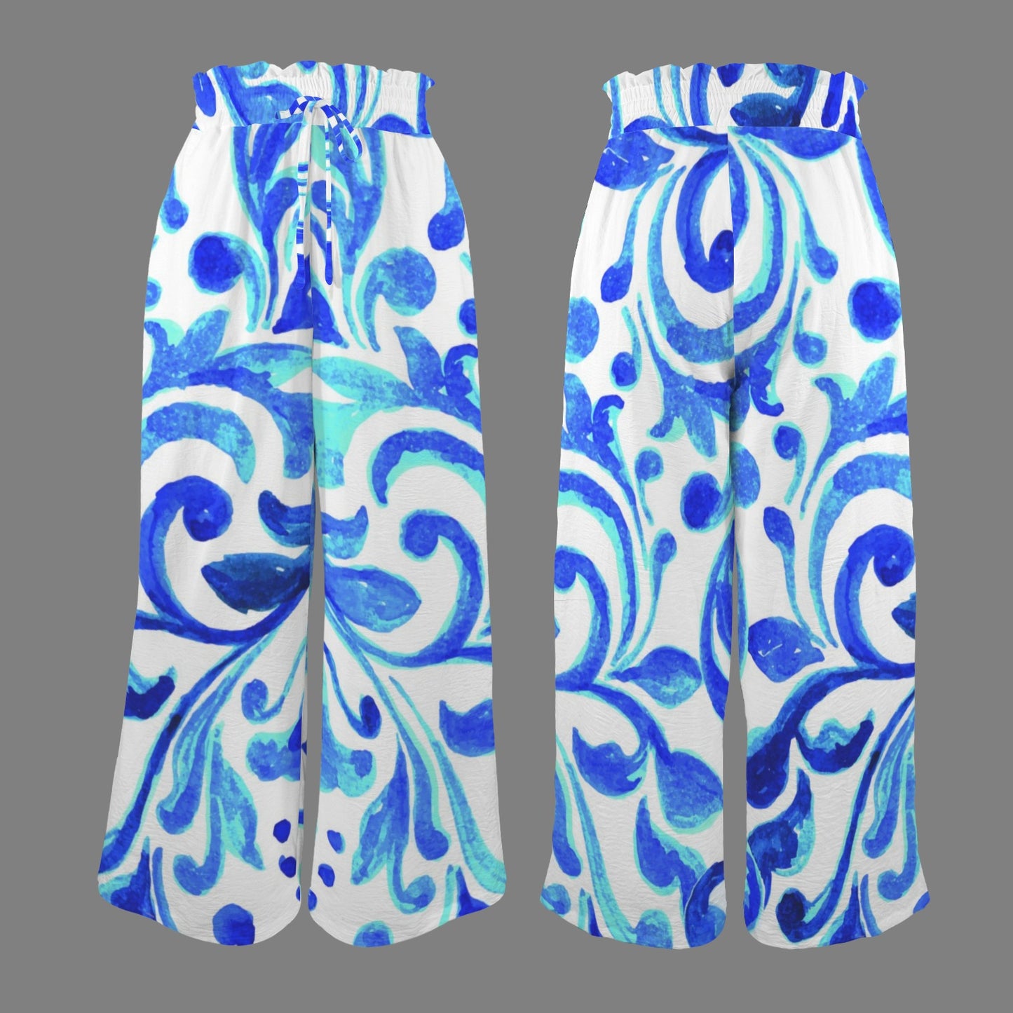 Full-body shot of a woman wearing Ondas Wide Leg Pants featuring a vibrant blue and white Mediterranean scroll print, paired with a white sleeveless top and straw purse, standing on a path at a tropical resort.