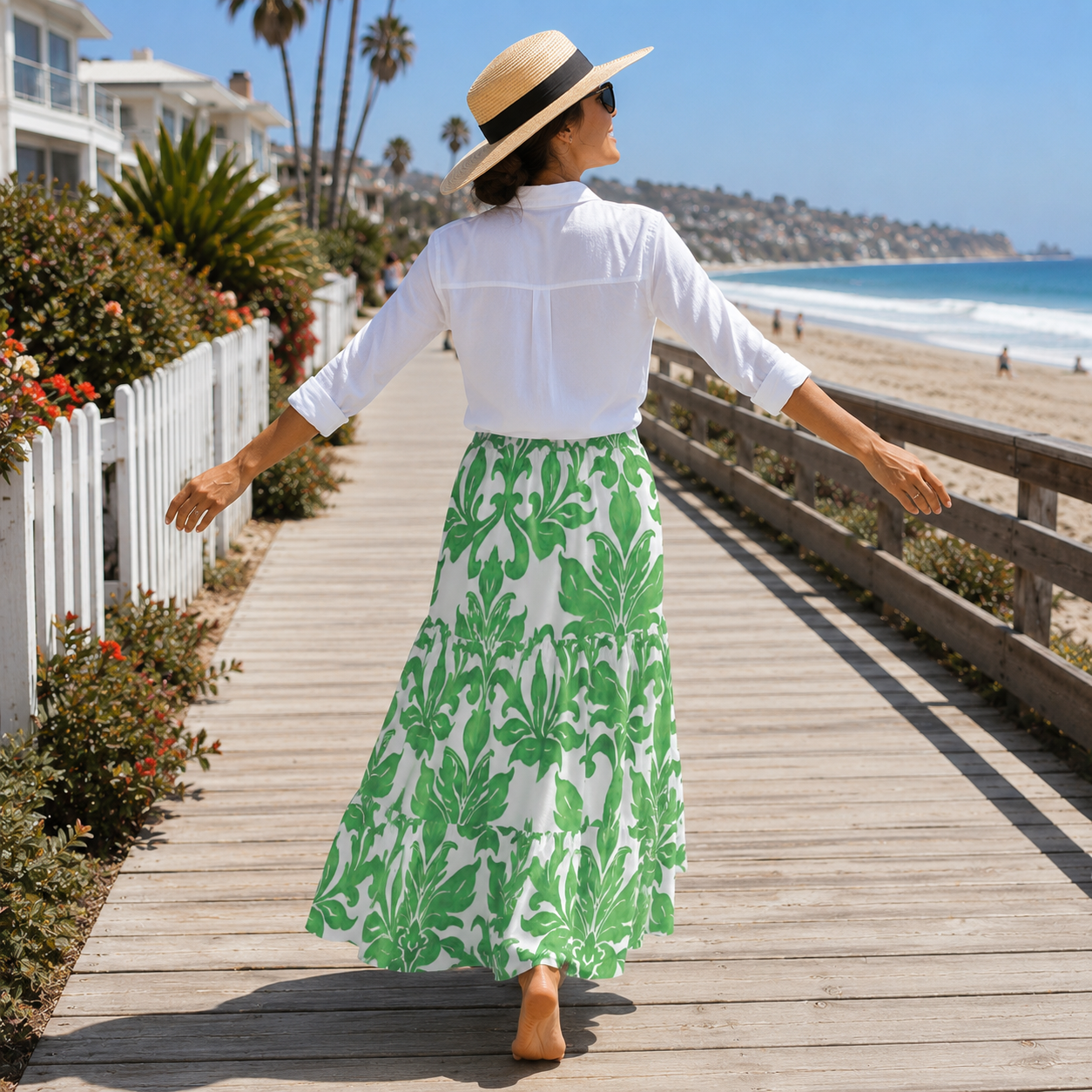 A woman viewed from behind walking on a wooden boardwalk by the beach, wearing a white long-sleeve blouse and a white maxi skirt with a bold green botanical leaf pattern, along with a straw hat.