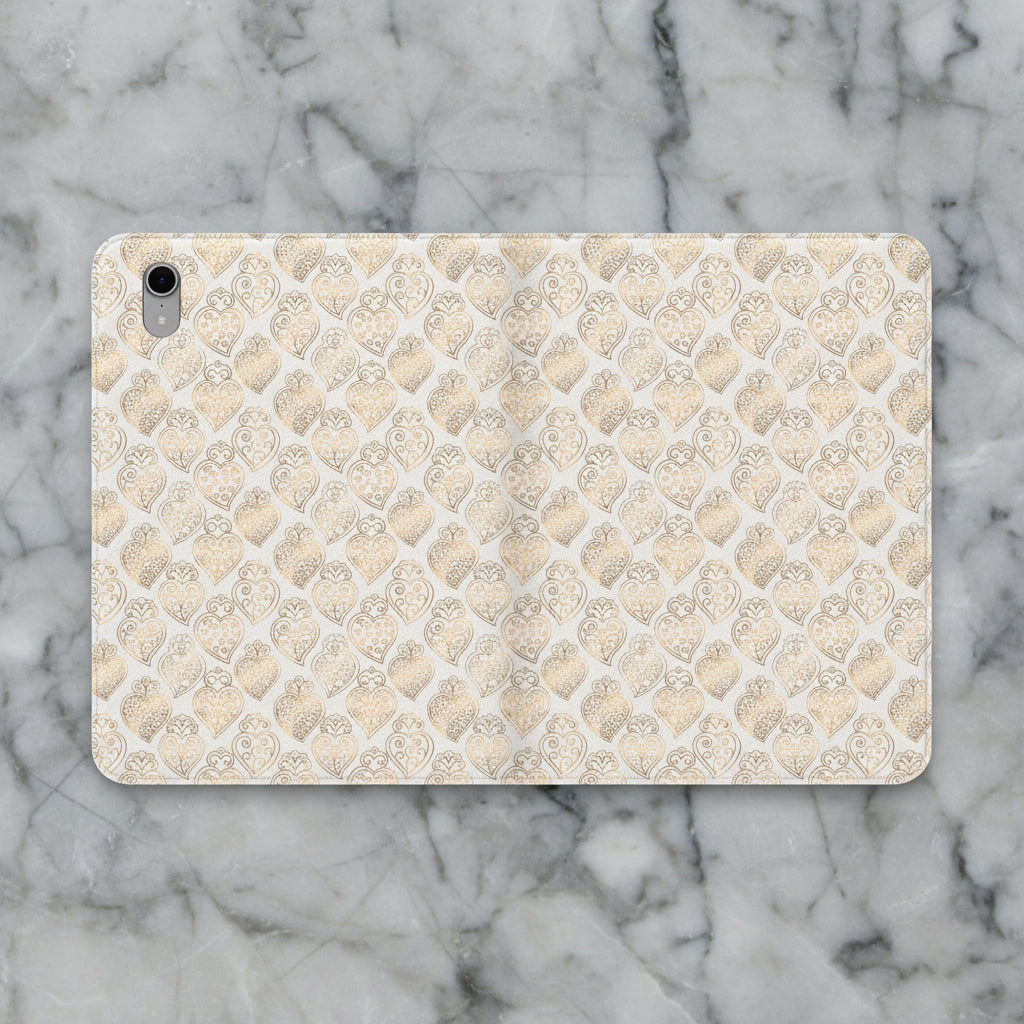 A protective tablet case featuring a repeating pattern of gold and silver filigree hearts on a light beige background, shown against a plain white studio backdrop.
