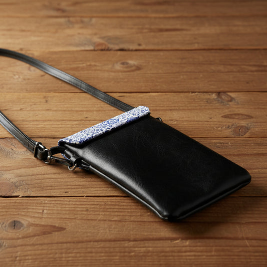 A black faux-leather crossbody phone bag with a decorative blue and white patterned fabric trim across the top, shown against a wood table