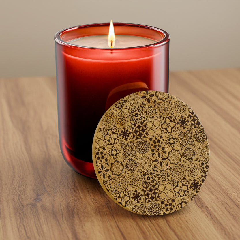 A lit red glass candle jar with a single flame, shown beside a round wooden lid or coaster featuring an intricate dark brown geometric and floral pattern, on a clean white background.