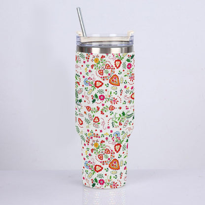 A tall insulated tumbler with a clear lid and metal straw, covered in a colorful floral and heart pattern on a white background, photographed against a light gray studio backdrop.