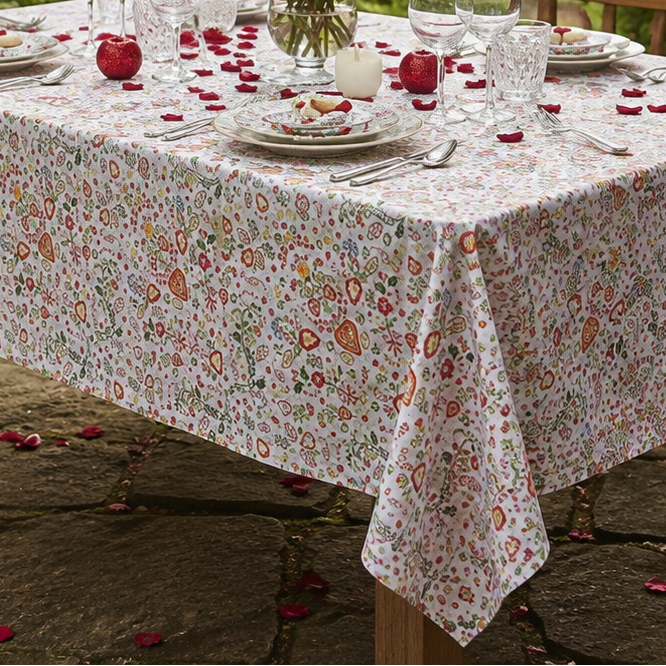 A rectangular table covered with a white tablecloth featuring a dense small floral print in muted red, pink, and green, set outdoors in a garden with place settings, wine glasses, a vase of red roses, and scattered rose petals.
