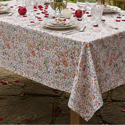A rectangular table covered with a white tablecloth featuring a dense small floral print in muted red, pink, and green, set outdoors in a garden with place settings, wine glasses, a vase of red roses, and scattered rose petals.