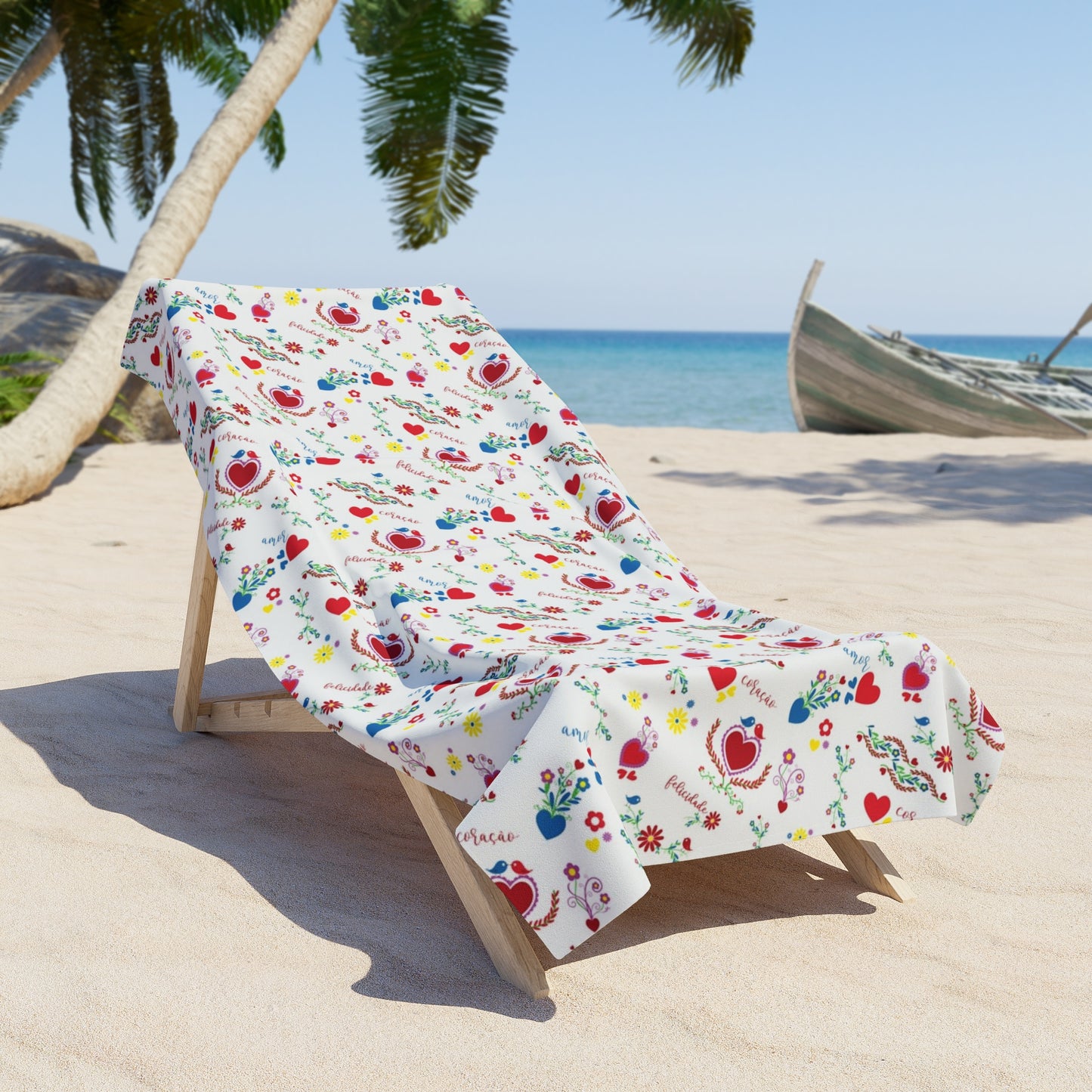A beach towel with a colorful floral pattern, laid out on the sand with a palm tree and part of a wooden beach chair visible in the background.