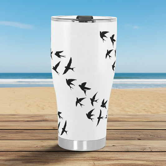 A white stainless steel tumbler with a pattern of black birds (sparrows) on a wooden surface with a sofa in the background.
