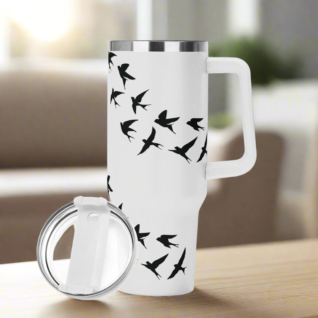 A white stainless steel tumbler with a handle and straw, decorated with a pattern of black swallows (andorinhas), placed on a wooden surface with a blurred background.