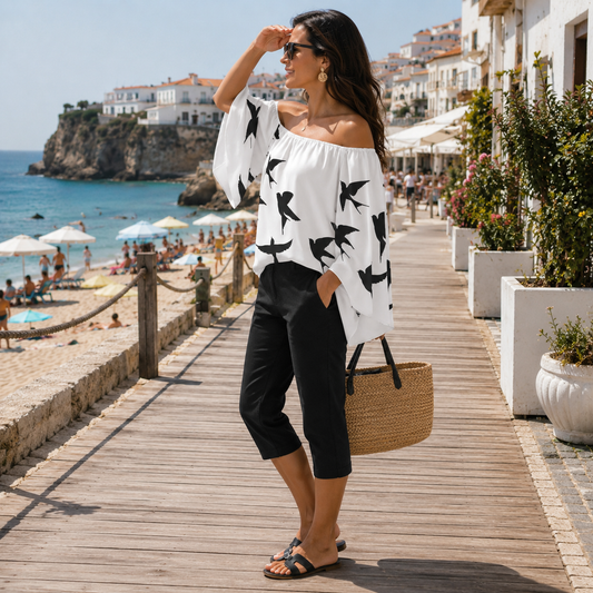 A woman stands on a wooden boardwalk by the beach wearing a white off-the-shoulder blouse with black bird prints, black cropped pants, and carrying a woven straw tote bag.