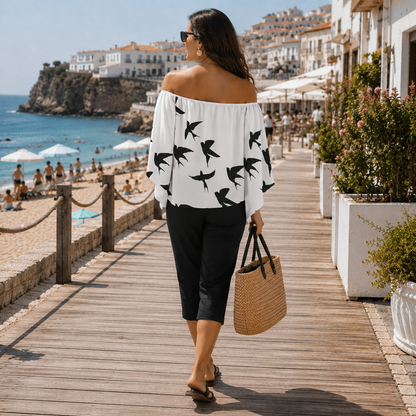 A woman walking on a wooden boardwalk by the sea, wearing an off-the-shoulder white top with black bird silhouettes, black cropped pants, and carrying a woven straw tote bag.