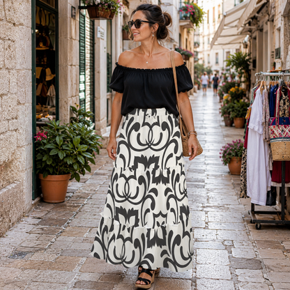 A woman walking down a cobblestone street wearing a black off-the-shoulder short-sleeve top and a long white maxi skirt with a bold black damask pattern. She is wearing sunglasses, a small shoulder bag, and black sandals.