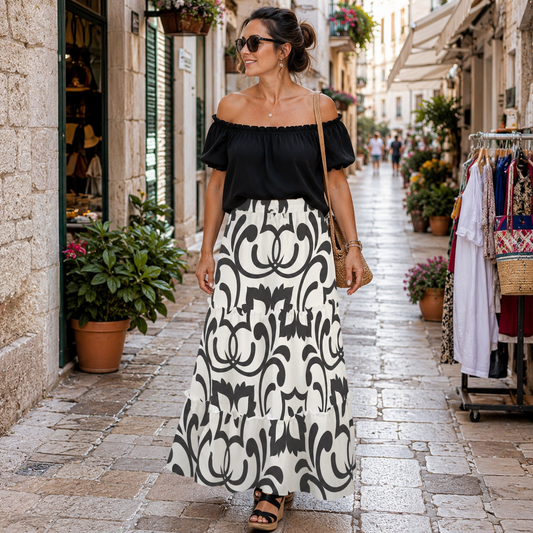 A woman walking down a cobblestone street wearing a black off-the-shoulder short-sleeve top and a long white maxi skirt with a bold black damask pattern. She is wearing sunglasses, a small shoulder bag, and black sandals.