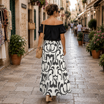 A woman viewed from behind wearing a black off-the-shoulder short-sleeve top and a long white maxi skirt with a bold black baroque scroll pattern, walking down a cobblestone street in a European town.