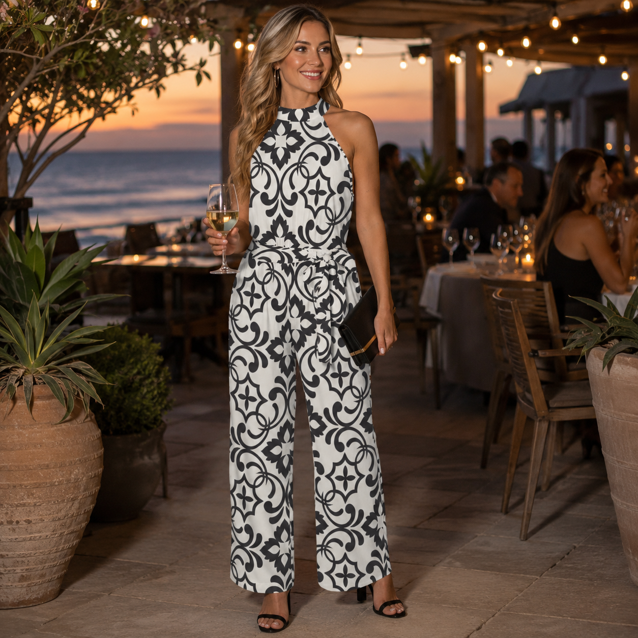 A woman stands at an outdoor evening patio by the ocean, wearing a sleeveless white jumpsuit with a bold black damask-style pattern and holding a glass of white wine.
