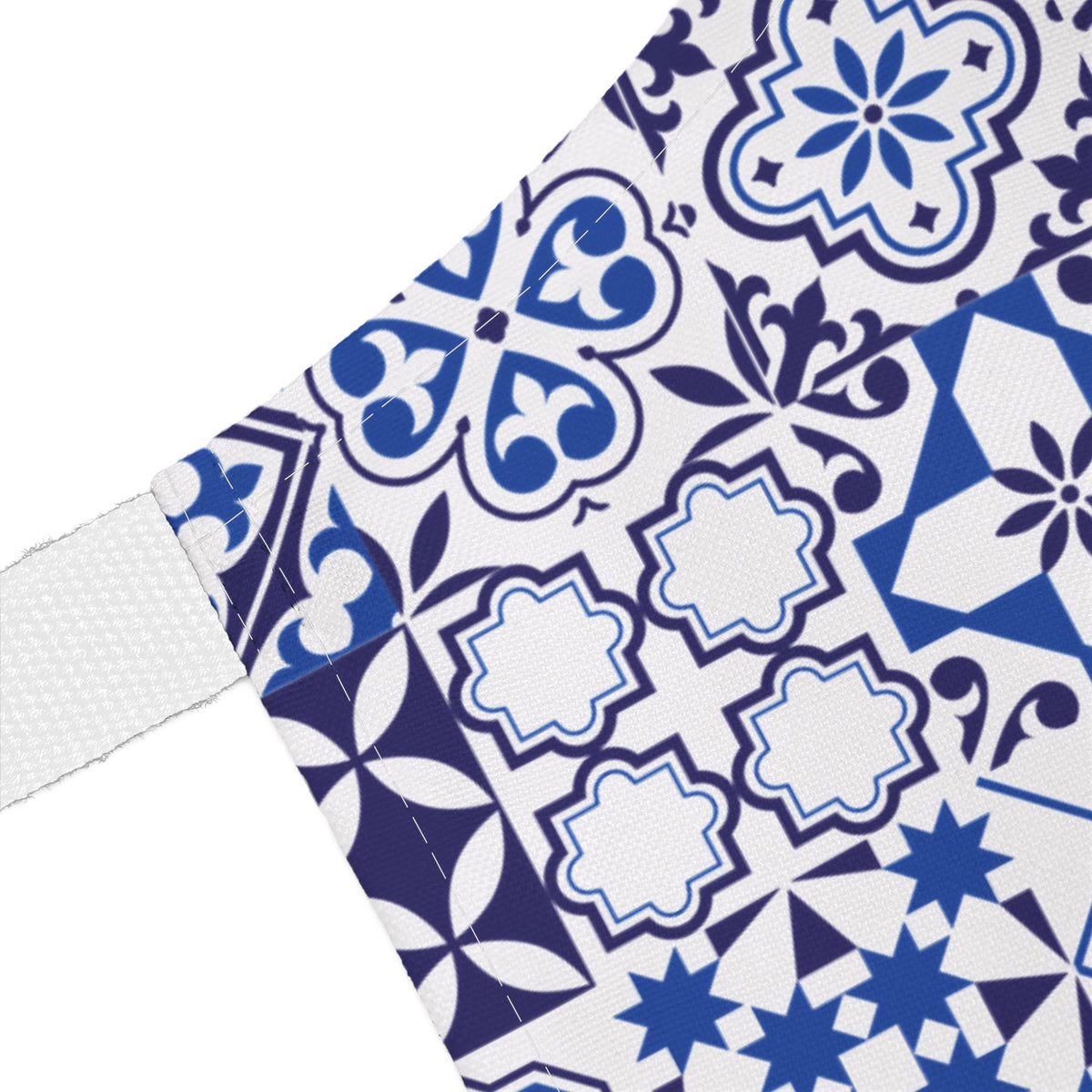 A close-up of a white fabric item, likely a towel or blanket, featuring a blue and navy Portuguese tile pattern. A white woven strap is visible on the left side against a plain white background.