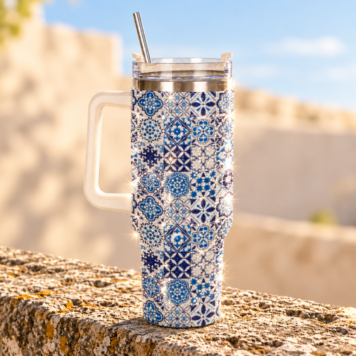A handled travel tumbler with a blue and white geometric tile pattern, a clear lid, and a metal straw, photographed outdoors on a stone surface with a blurred sunny background.