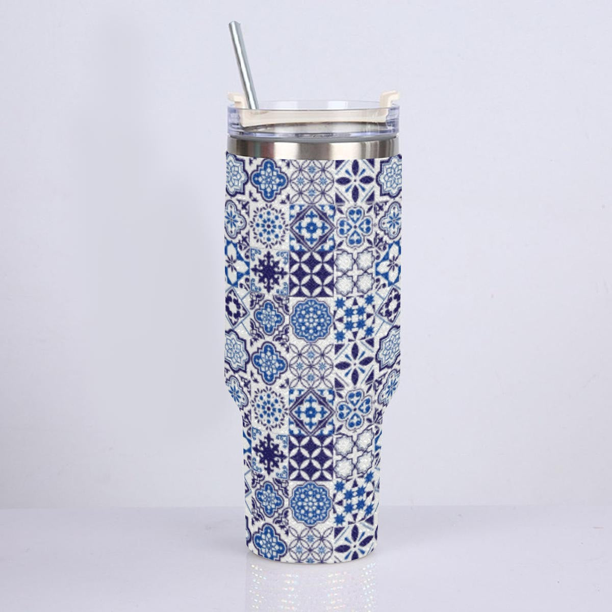 A tall insulated tumbler with a blue and white geometric tile pattern wrap, featuring a clear lid with a silver metal straw, set against a plain light gray background.