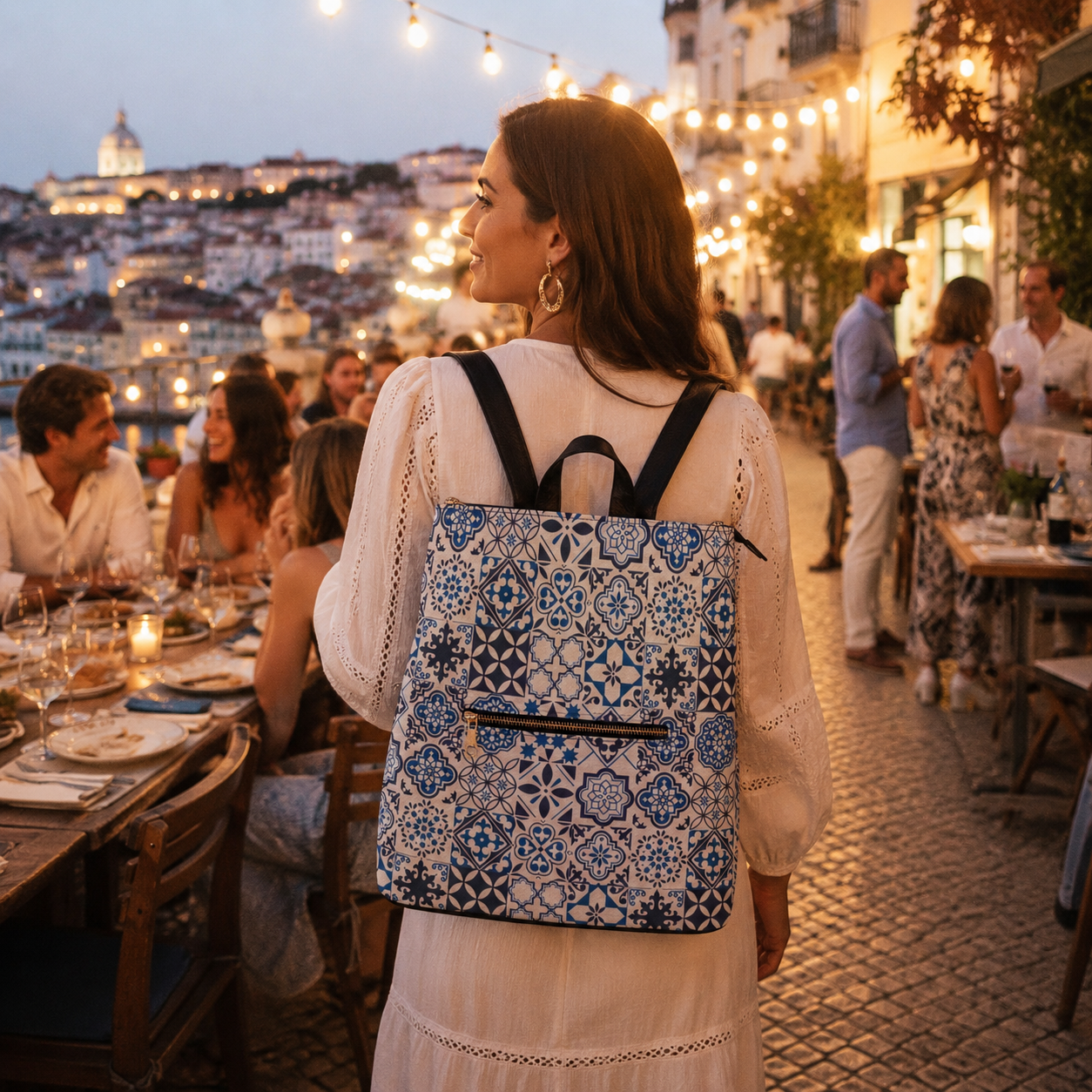 A woman seen from behind at an outdoor evening restaurant, wearing a blue and white patterned backpack with black shoulder straps. The background shows a lively dining scene with warm string lights and a scenic city view in the distance.