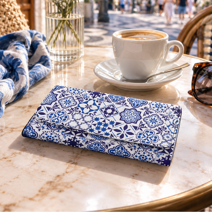 A blue and white patterned clutch wallet lies on a marble cafe table next to a white coffee cup, sunglasses, a woven bag, and a vase of flowers, with a blurred outdoor plaza in the background.