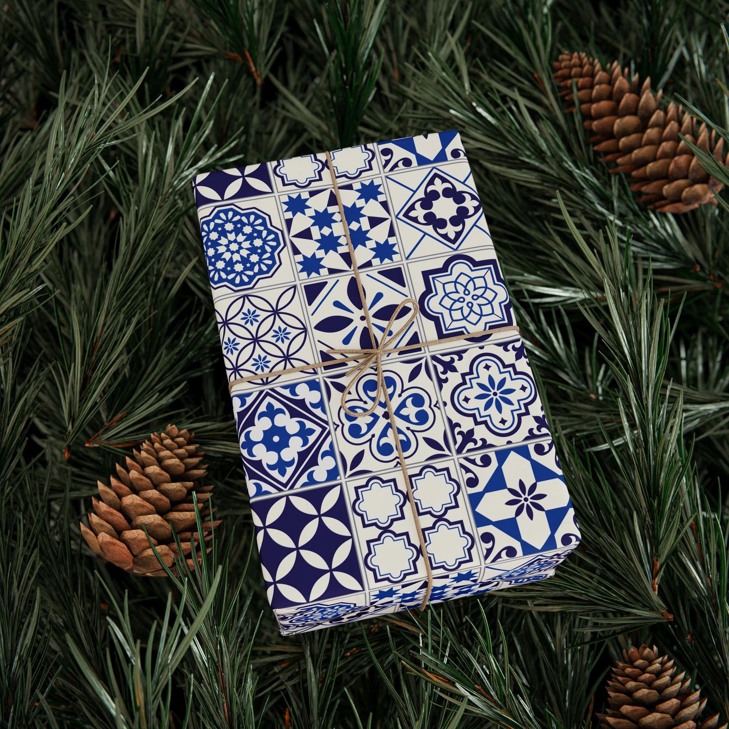 Gift box wrapped in blue and white tile patterned paper on green foliage with pine cones.