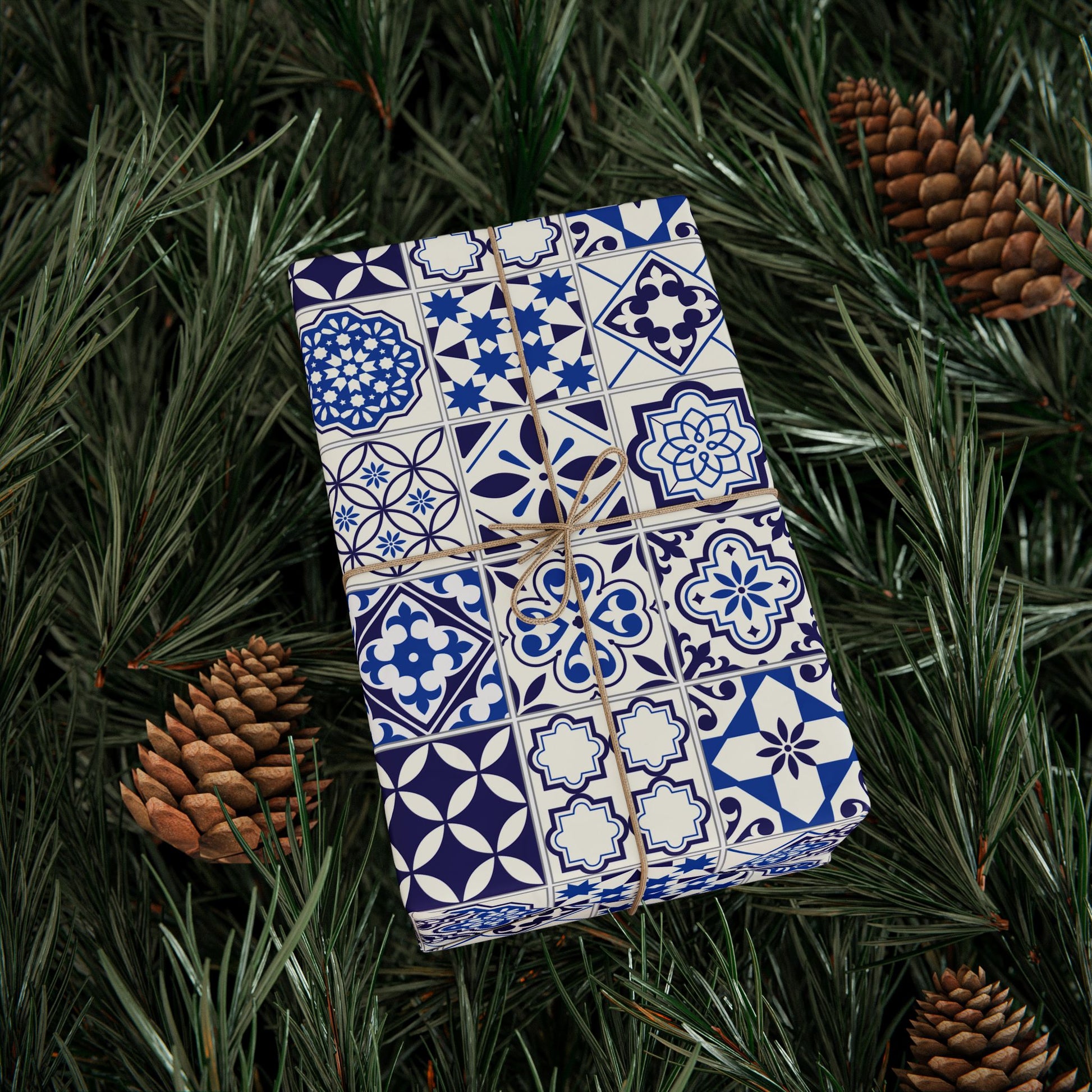 Gift box wrapped in blue and white tile patterned paper on green foliage with pine cones.