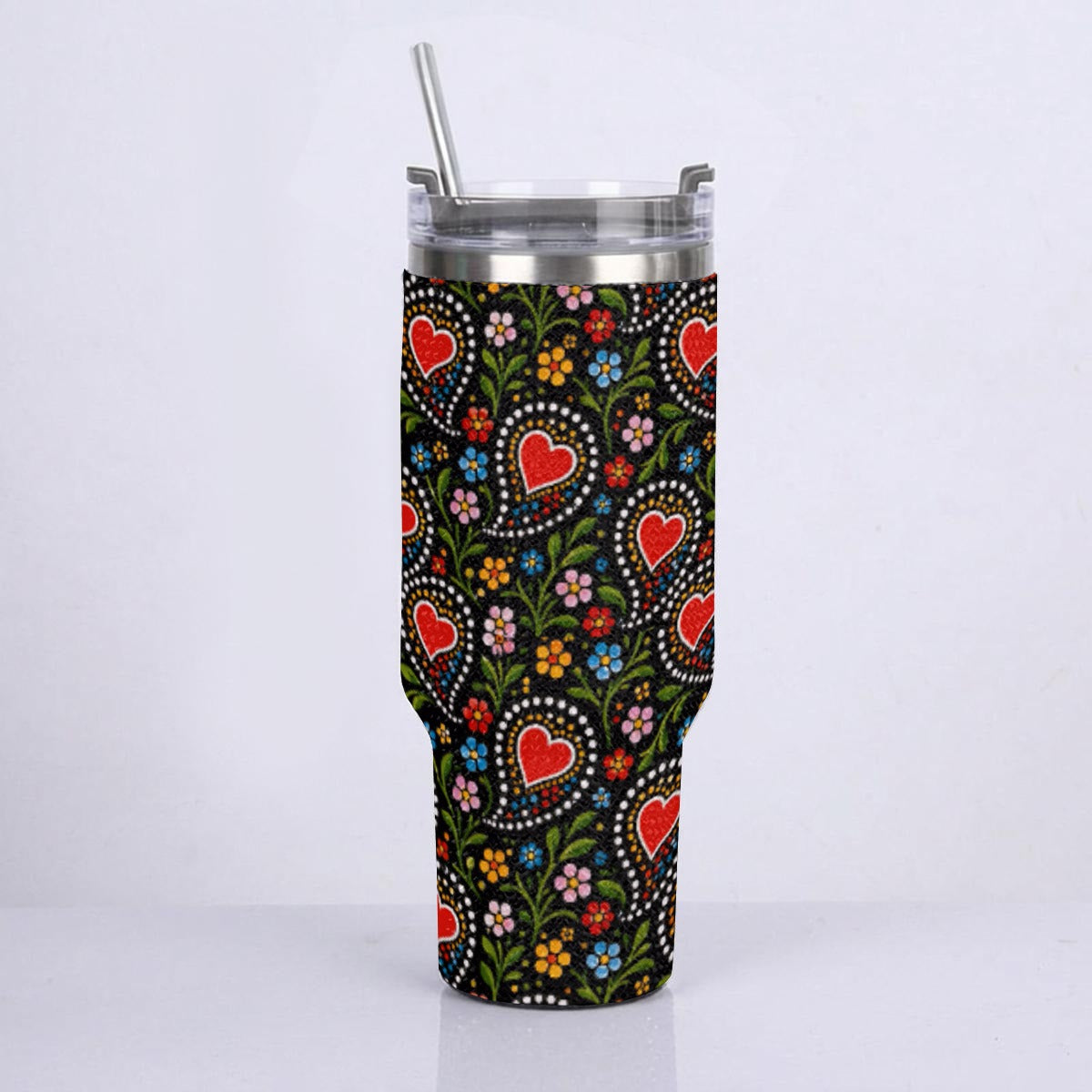 A tall insulated tumbler with a clear lid and metal straw, covered in a black sleeve featuring a colorful floral and heart pattern, shown on a light gray background.