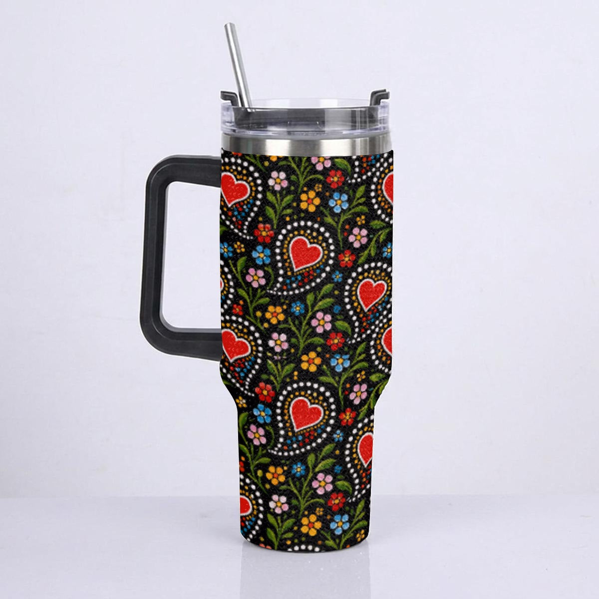 A handled travel tumbler with a clear lid and metal straw, featuring a black background covered in a colorful floral and heart pattern, shown against a plain white studio background.