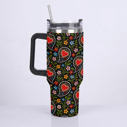 A handled travel tumbler with a clear lid and metal straw, featuring a black background covered in a colorful floral and heart pattern, shown against a plain white studio background.