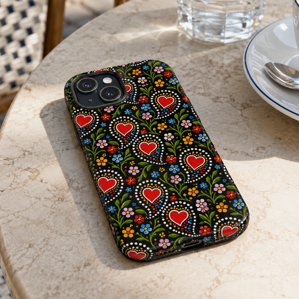 A smartphone case with a colorful folk-art style pattern of red hearts, green leaves, and small flowers on a black background, shown resting on a round stone table next to a glass of water and a cup of coffee.