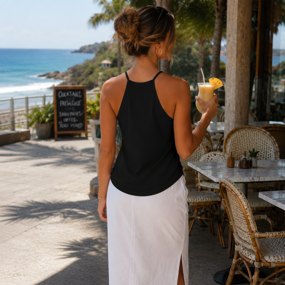A woman stands on a beachside patio wearing a black halter-neck tank top and a white midi skirt with a side slit. She is holding a drink with a pineapple slice garnish.