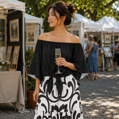 A woman stands outdoors at an art fair wearing an off-the-shoulder black blouse and a long black-and-white patterned skirt, holding a glass of champagne. Paintings on easels and white tents are visible in the background.
