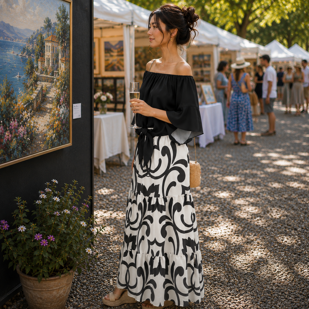 A woman stands outdoors at an art fair or market, wearing an off-the-shoulder black top and a long black-and-white patterned skirt. She holds a drink and looks to the side, with framed paintings, white tents, trees, and other people in the background.