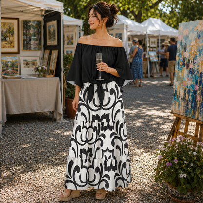 A woman stands outdoors at an art fair wearing an off-the-shoulder black blouse and a long black-and-white patterned skirt, holding a glass of champagne. Paintings on easels and white tents are visible in the background.
