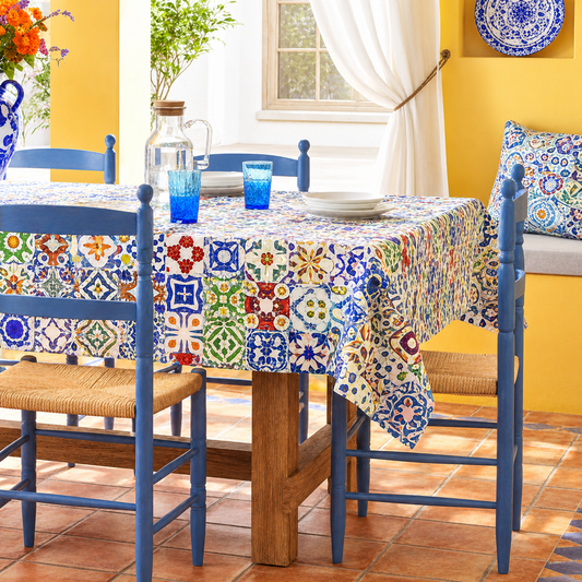 A colorful patchwork tile-patterned tablecloth draped over a wooden dining table, surrounded by blue chairs in a bright yellow room.