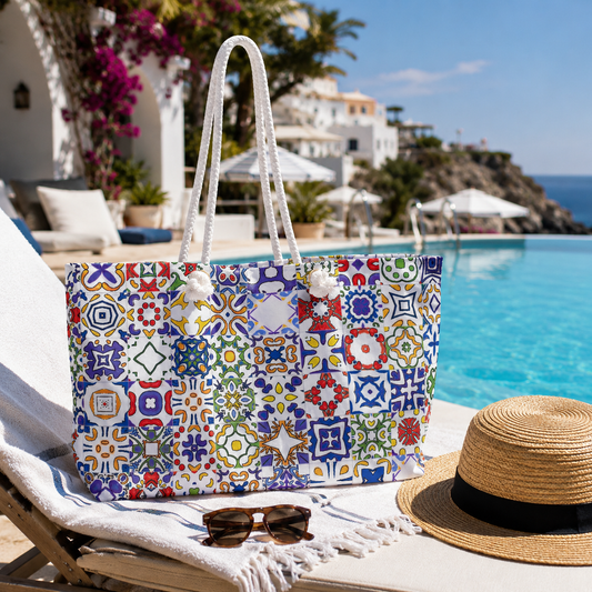 A large tote bag with a colorful Mediterranean tile pattern sits on a white lounge chair next to a swimming pool. A straw sun hat with a black band and a pair of sunglasses are placed beside it, with a white villa and blue sea in the background.