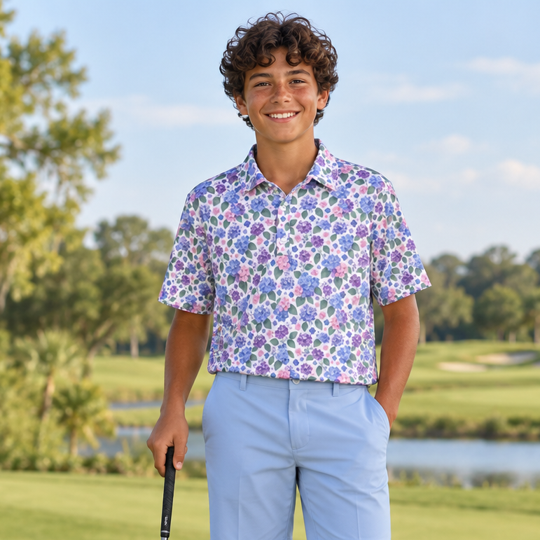 A young man stands on a golf course wearing a short-sleeve button-up shirt with a colorful floral pattern, light blue shorts, and white golf shoes, holding a driver. A golf bag is visible on the left, with trees and water in the background.
