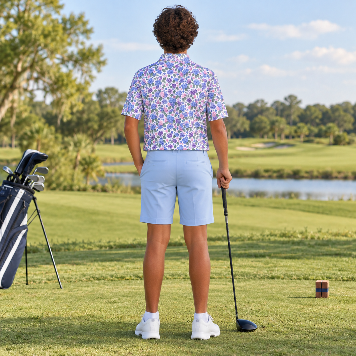 A man on a golf course seen from behind, wearing a short-sleeve button-up shirt with a blue, purple, and white floral pattern, light blue shorts, and white golf shoes. He is holding a golf club, with a golf bag and tee visible in the background.