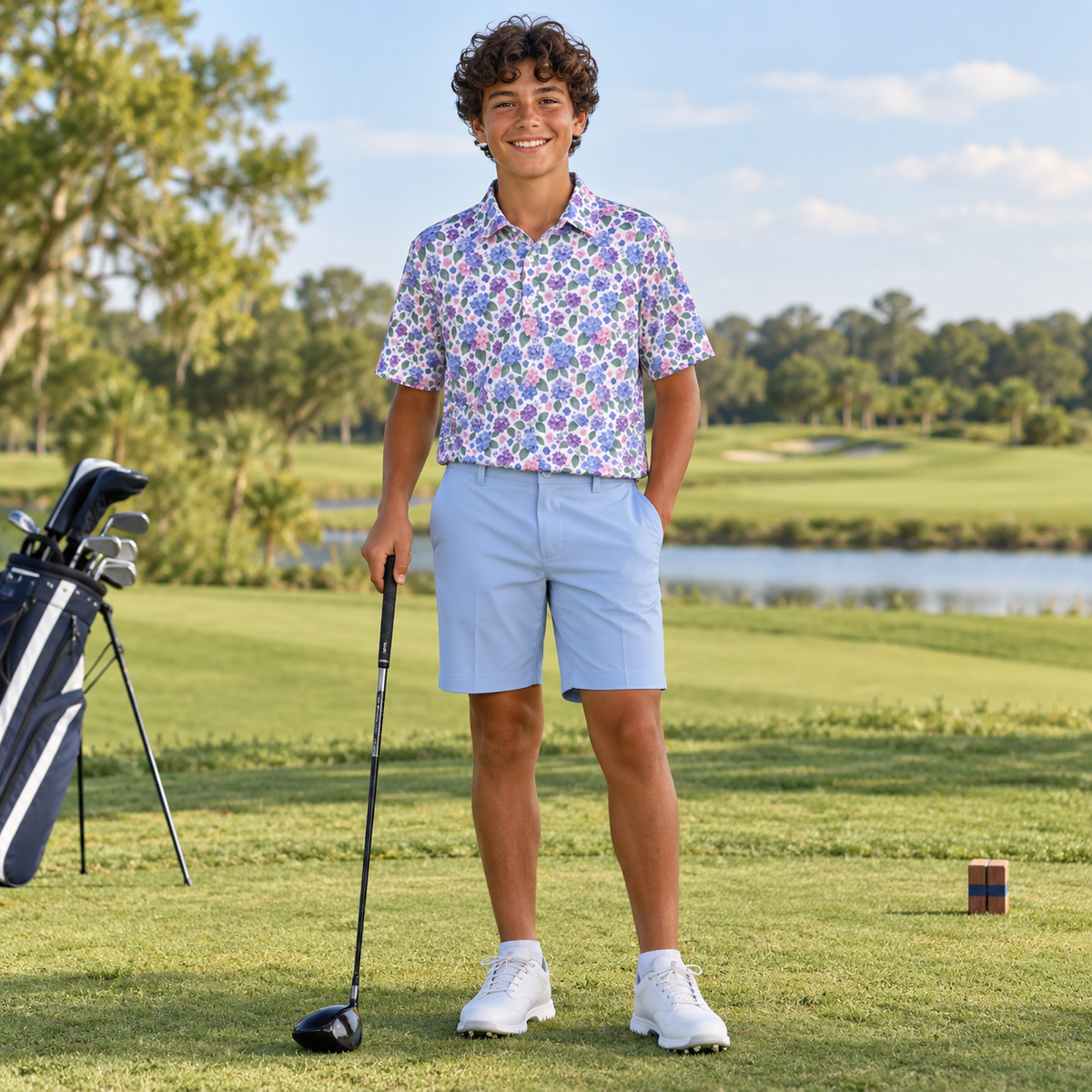 A young man stands on a golf course wearing a short-sleeve button-up shirt with a colorful floral pattern, light blue shorts, and white golf shoes, holding a driver. A golf bag is visible to his left.