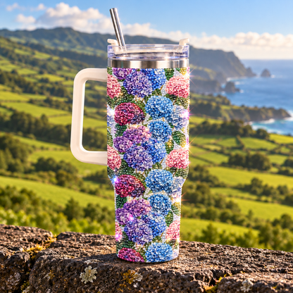 A handleless travel tumbler with a clear lid and metal straw, covered in a dense floral bead pattern of pink, purple, and blue hydrangeas, set against a scenic coastal mountain background.