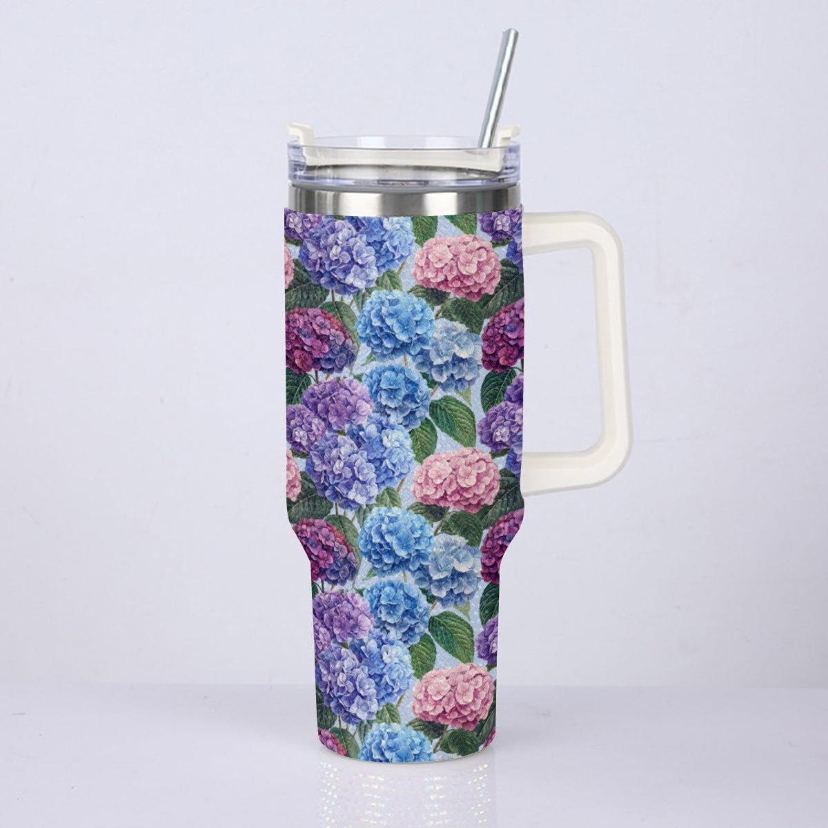 A tall insulated tumbler with a floral print featuring blue, pink, and purple hydrangeas with green leaves. The tumbler has a white handle, a clear lid with a metal straw, and a silver stainless steel rim, shown against a plain light gray background.