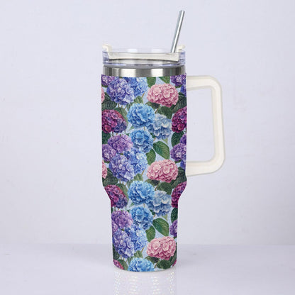 A tall insulated tumbler with a floral print featuring blue, pink, and purple hydrangeas with green leaves. The tumbler has a white handle, a clear lid with a metal straw, and a silver stainless steel rim, shown against a plain light gray background.