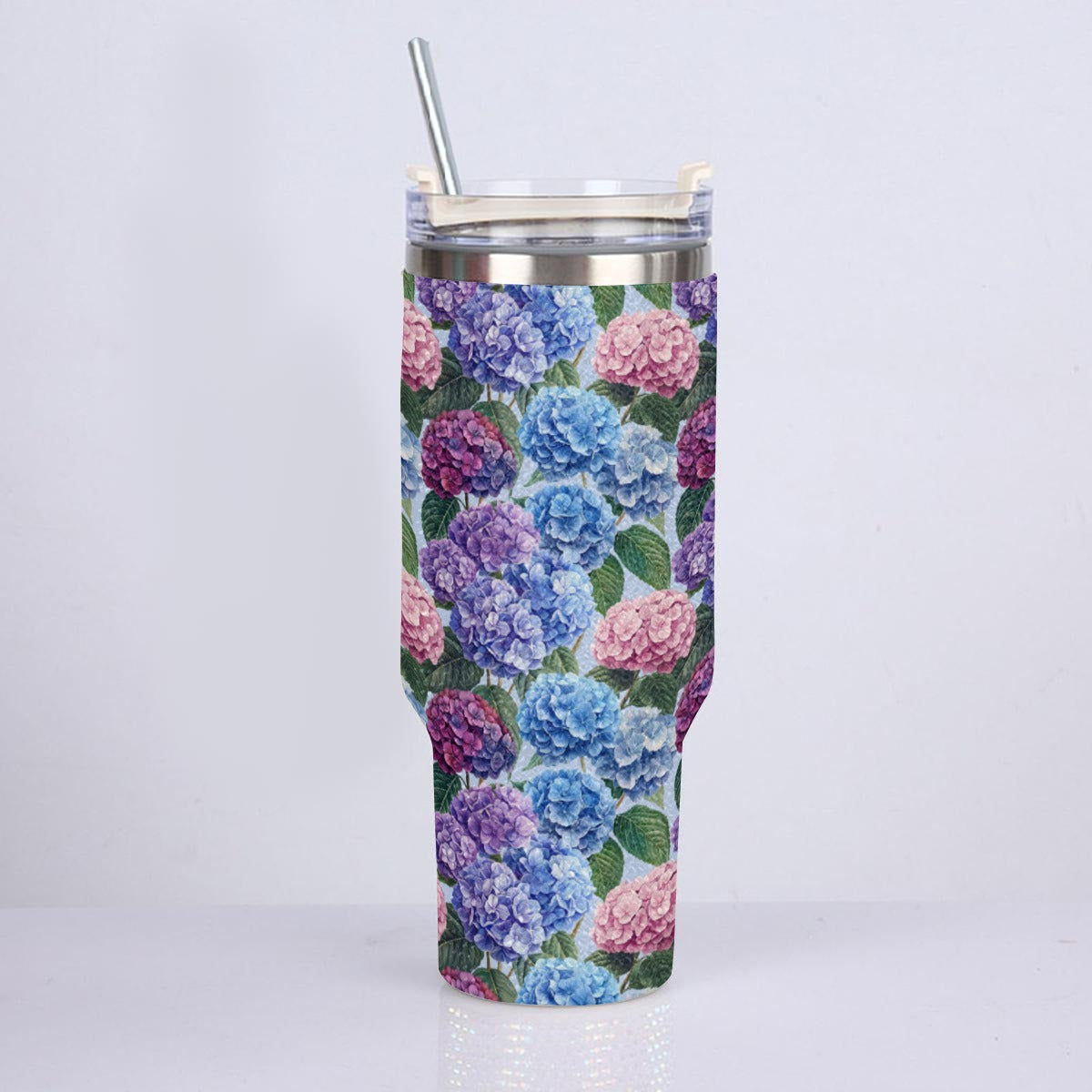 A tall insulated tumbler with a floral print featuring blue, pink, and purple hydrangeas on a dark background, shown with a clear lid and metal straw against a plain light gray background.