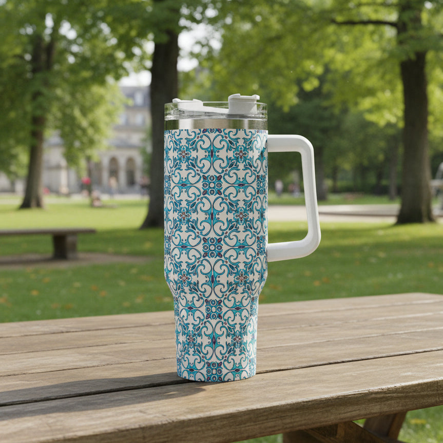 A 40oz stainless steel tumbler with a Portuguese tile-inspired design, featuring a handle and straw, held in a hand with a background of colorful flowers.