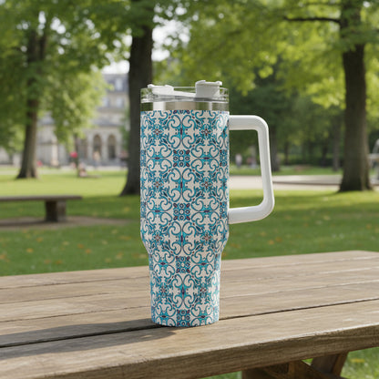 A 40oz stainless steel tumbler with a Portuguese tile-inspired design, featuring a handle and straw, held in a hand with a background of colorful flowers.