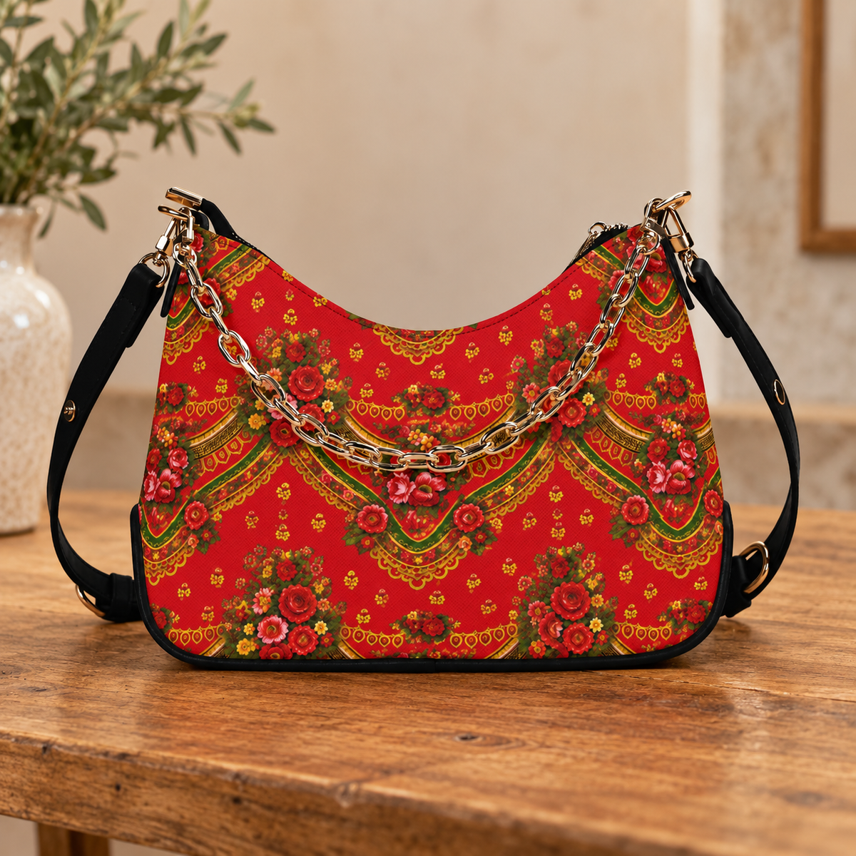 A red shoulder bag with a traditional floral and geometric pattern, featuring a gold chain strap and black shoulder straps, displayed on a wooden table.