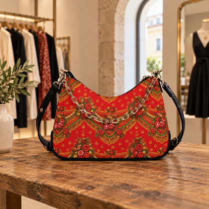 A red shoulder bag with an ornate floral and geometric pattern, featuring a gold chain strap and black leather straps, displayed on a rustic wooden table inside a boutique.