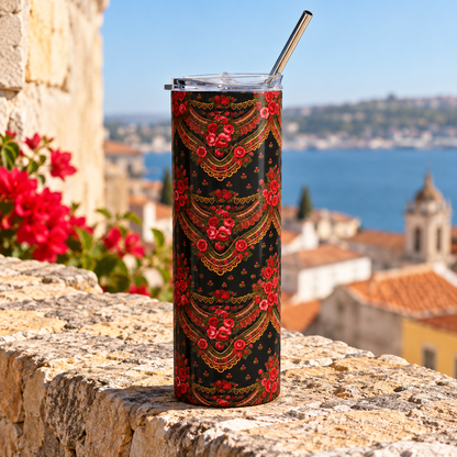 A tall insulated tumbler with a clear lid and metal straw, featuring a black background with a red and gold ornate floral pattern, set on a stone ledge with a coastal town and sea in the background.