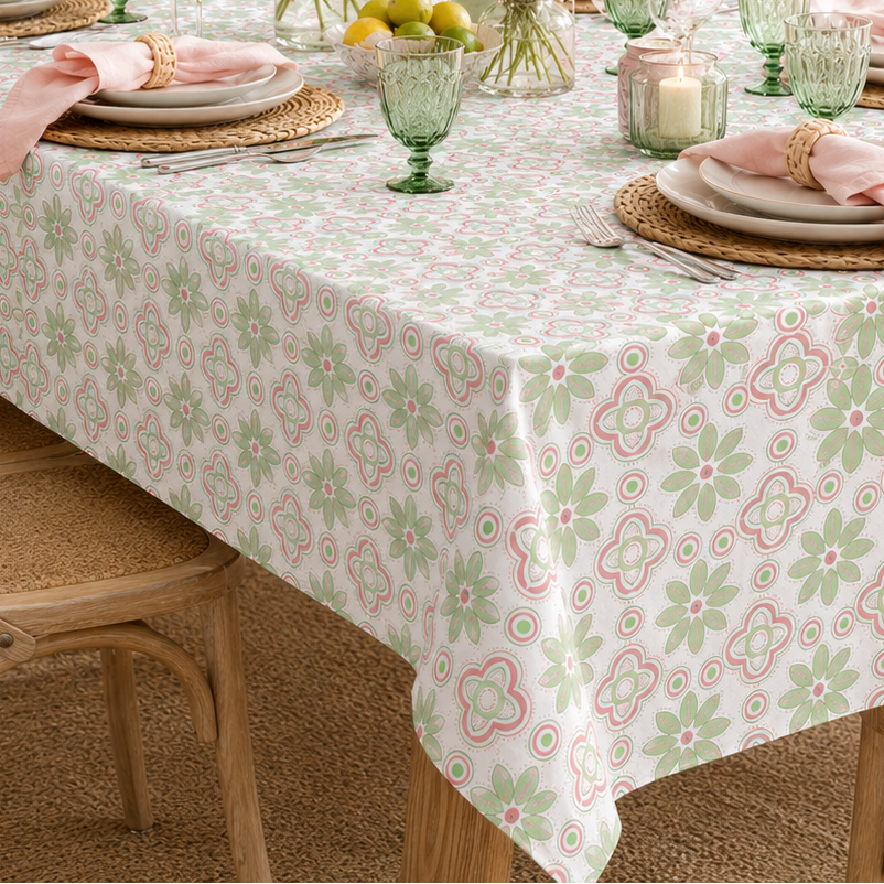 A rectangular dining table covered with a white tablecloth featuring a repeating pastel floral pattern in pink and sage green. The table is set with plates, pink napkins, green glassware, woven placemats, and multiple floral arrangements, surrounded by wooden chairs in a bright, sunlit dining room.