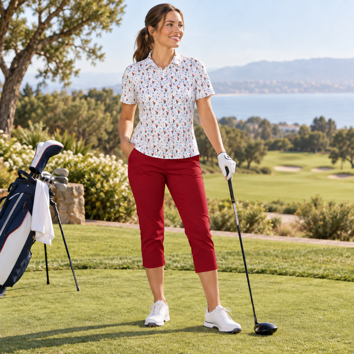 A woman stands on a golf course wearing a short-sleeve white collared shirt with a small multicolor floral print, burgundy cropped pants, white golf shoes, and a white glove. She holds a golf club, with a golf bag beside her.