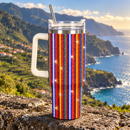 A handleless insulated tumbler with a white handle and metal straw, covered in vertical red, white, and blue rhinestone stripes, set against a scenic coastal mountain background.